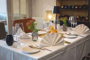 Festlich eingedeckte Tafel mit weißer Tischdecke, gefalteten Servietten und Weinregal im Hintergrund